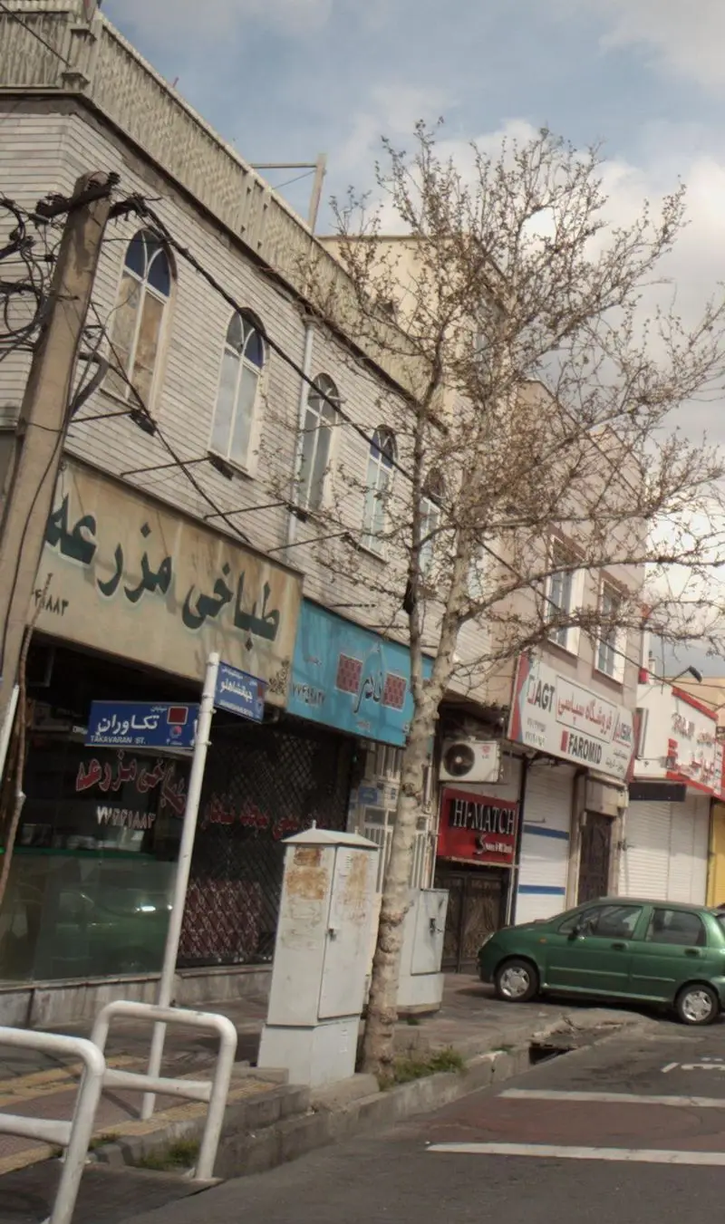 تصویری از نمای خیابان و مغازه طباخی مزرعه با تابلوی بزرگ و واضح، در کنار سایر فروشگاهها و یک ماشین سبز رنگ پارک شده.