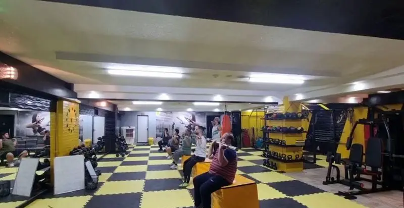 باشگاه ورزشی گلایل: محیطی مدرن و مجهز با کف‌پوش مشکی-زرد، دستگاه‌های بدنسازی متنوع، هالتر و دمبل. افراد در حال تمرینات ورزشی و تناسب اندام.