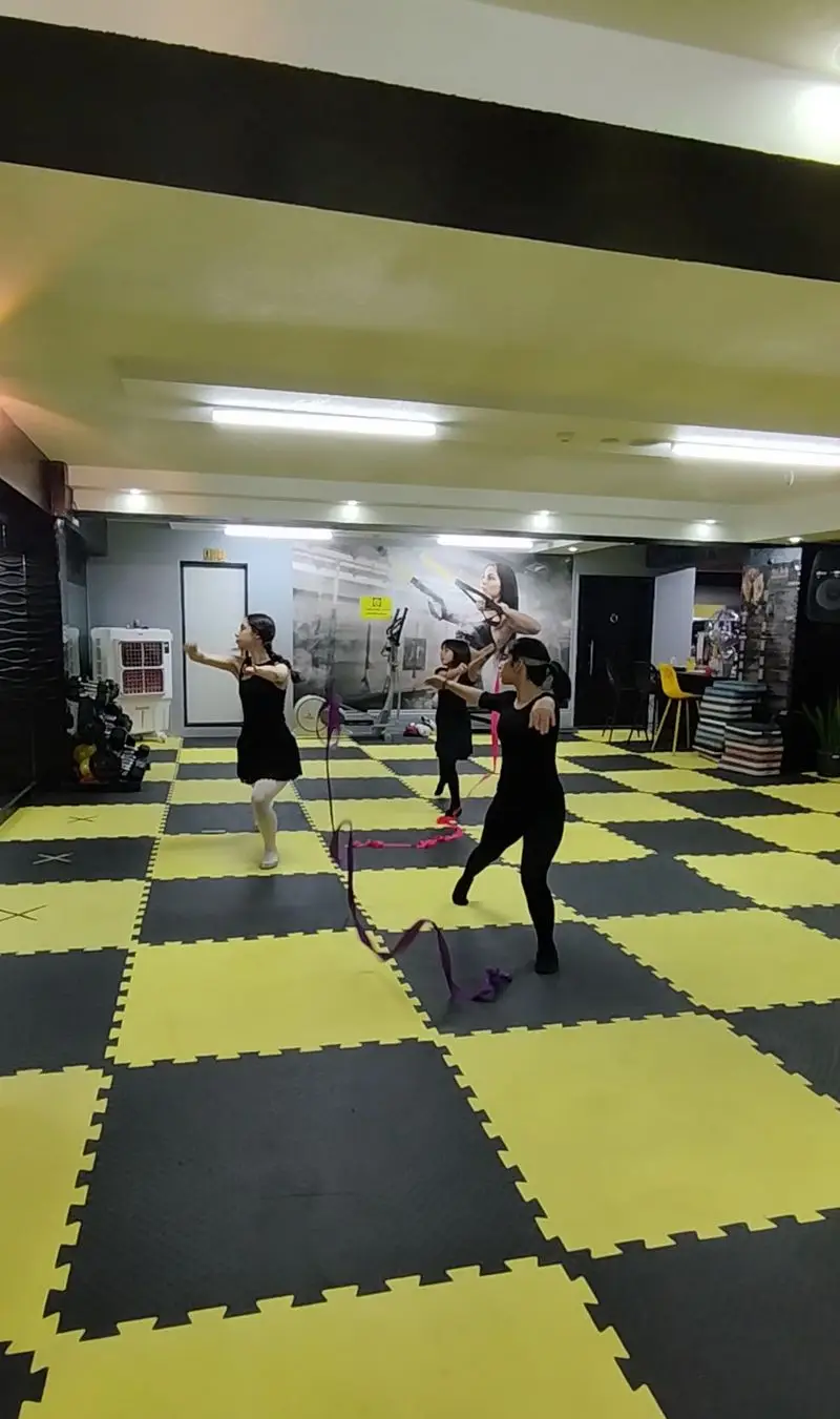 زنان در حال تمرین رقص با روبان در باشگاه ورزشی گلایل. کلاس ژیمناستیک ریتمیک با کف‌پوش شطرنجی و دیوارنگاره ورزشی در پس‌زمینه.