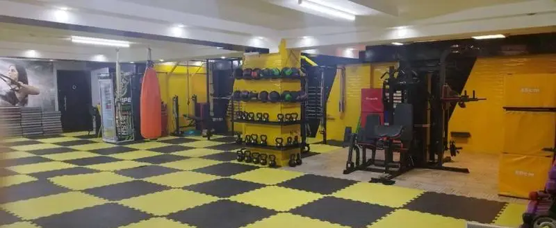 فضای داخلی باشگاه ورزشی گلایل: سالنی بزرگ با کفپوش شطرنجی زرد و مشکی، کیسه بوکس، کتل بل و دستگاه‌های بدنسازی پیشرفته.