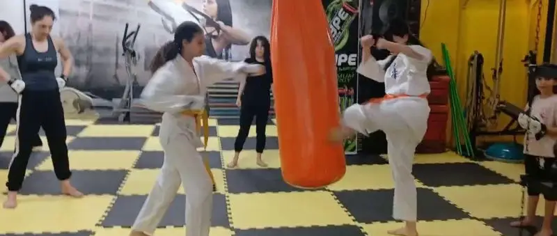 بانوان در حال تمرین هنرهای رزمی و کیک بوکسینگ در باشگاه ورزشی گلایل، شامل ضربه به کیسه بوکس و حرکات رزمی. محیط باشگاه ورزشی.