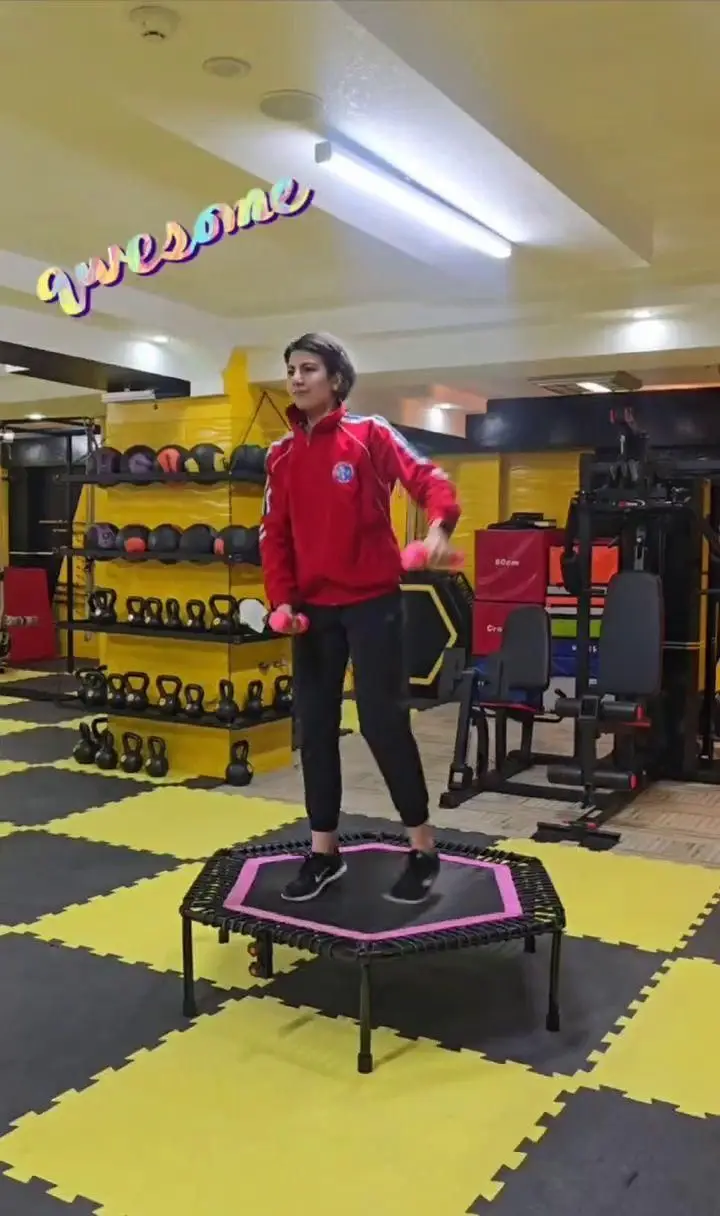 زنی پر انرژی در حال تمرین با ترامپولین و دمبل‌های صورتی در باشگاه ورزشی گلایل. فضایی مدرن و پویا برای تناسب اندام و سلامت بانوان.