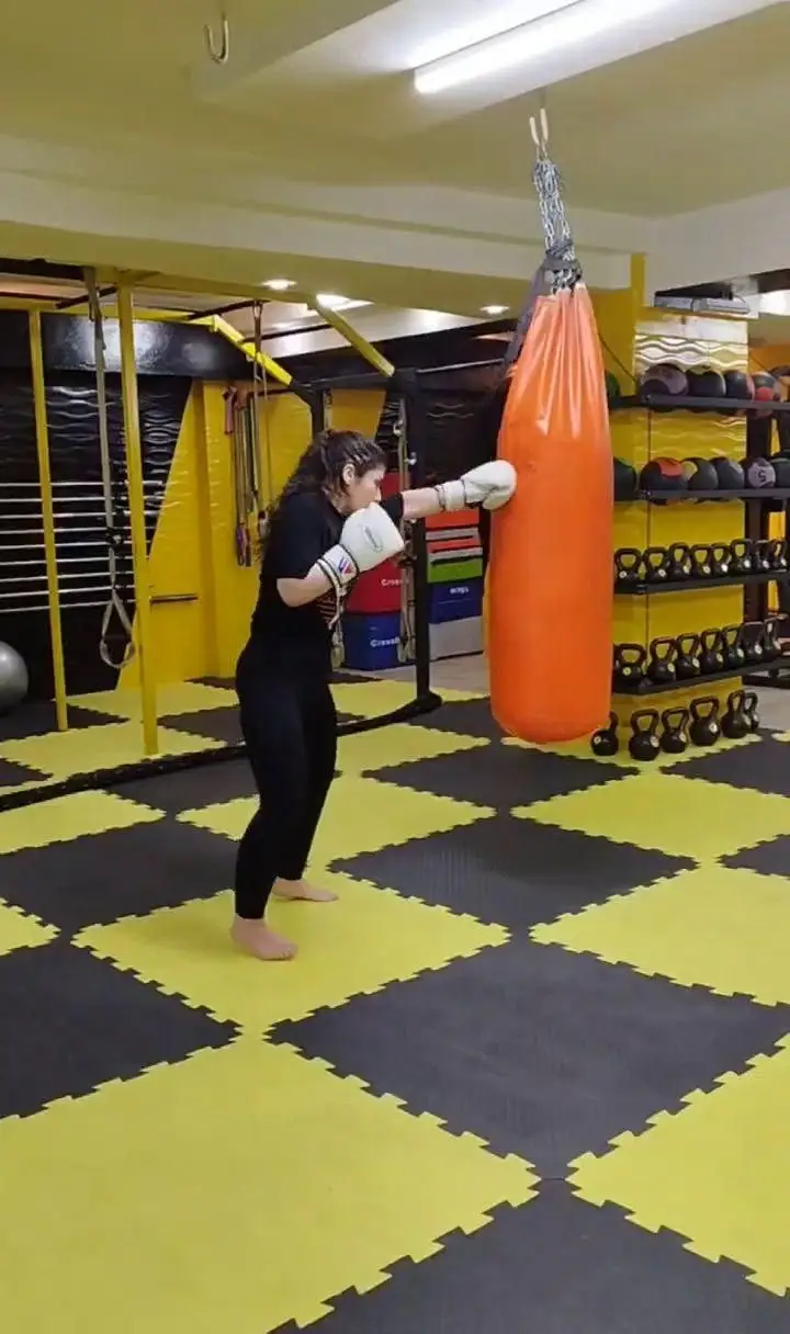 تصویر زنی در باشگاه ورزشی گلایل در حال تمرین بوکس با کیسه، نشانگر فضای فعال و تجهیزات کامل باشگاه برای تناسب اندام و ورزش.