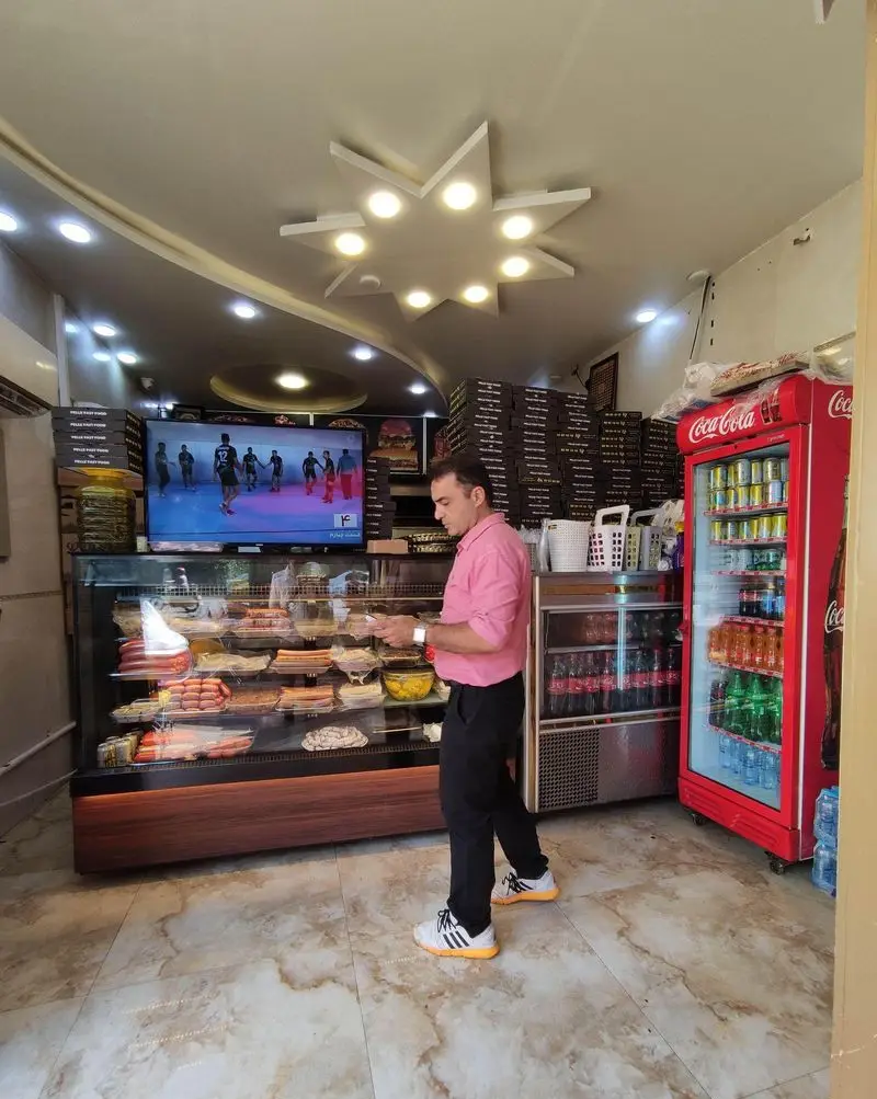 کارمند فست فود پله در حال سرویس‌دهی کنار ویترین غذا. نمایش سوسیس، کالباس، پیتزا و نوشیدنی‌های متنوع در یک فضای داخلی مدرن.