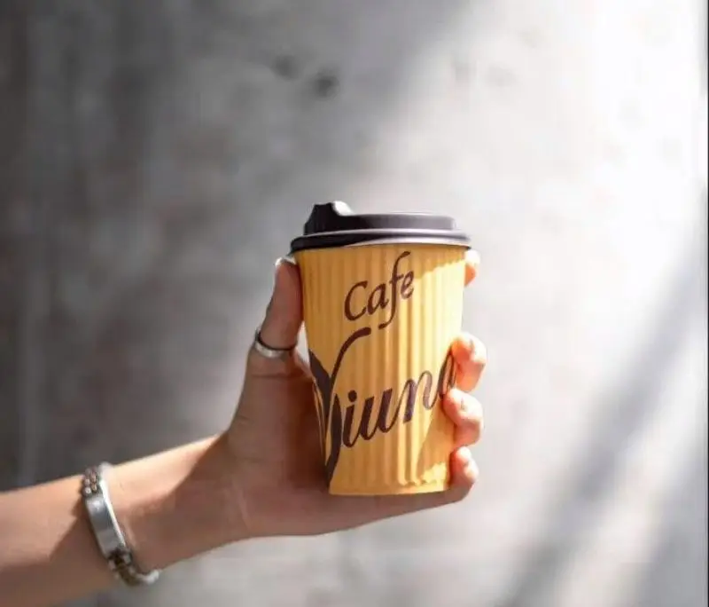 نمایی نزدیک از دست در حال نگه داشتن لیوان قهوه کاغذی زردرنگ با لوگوی 'کافه ویونا' برای لذت بردن از یک نوشیدنی گرم در لحظات دلپذیر.