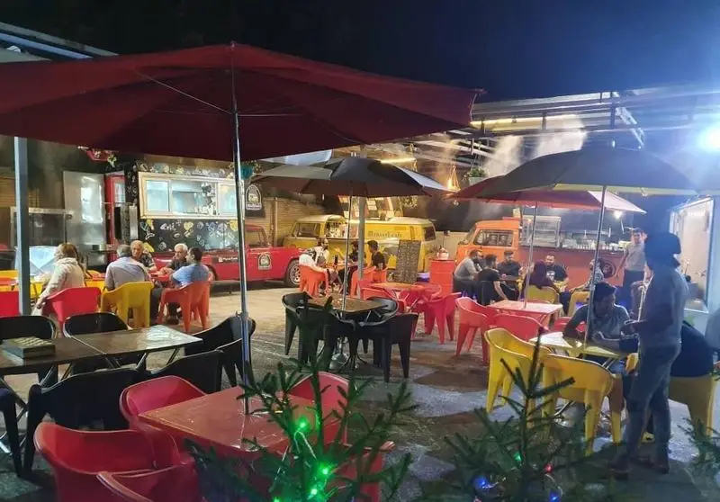 فضای شبانه کارفودلند با فودتراک‌های متنوع و افراد نشسته در میز و صندلی‌های رنگارنگ زیر چترها. محیطی شاد و پر جنب و جوش برای لذت از غذای خیابانی.