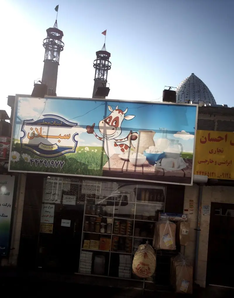 بیلبورد "لبنیات محلی سبلان" با گاو کارتونی و شیر، پنیر و ماست تازه، بر فراز مغازه‌ای با محصولات متنوع، در پس‌زمینه مناره‌ها.