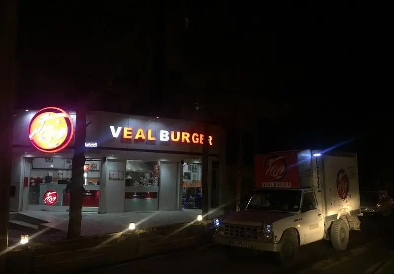 یک تصویر شب زیبا از فست فود ویل برگر، با تابلوی نئونی چشم‌نواز و لوگوی منحصربه‌فرد، کنار ون دلیوری آماده برای سرویس بیرون‌بر سریع.