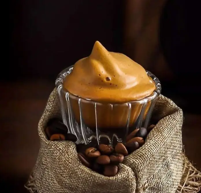 نوشیدنی فوم قهوه دالگونا غلیظ و جذاب در فنجان شیشه‌ای، در کنار دانه‌های قهوه بو داده شده، از محصولات ویژه قهوه یویو.