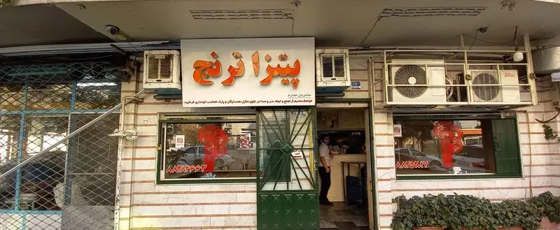تصویری از نمای بیرونی فست‌فود و پیتزا ترنج در روز، با تابلوی بزرگ و نارنجی "پیتزا ترنج" و ورودی مغازه. تهویه مطبوع نیز بالای آن دیده می شود.