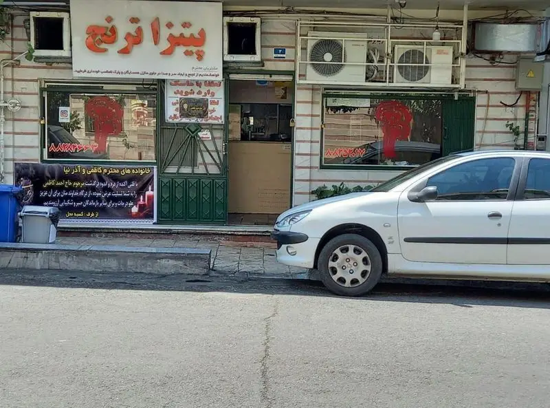 نمای خارجی فست‌ فود ترنج با تابلوی "پیتزا ترنج"، درب ورودی و بنر تسلیت، و یک خودروی سفید پژو ۲۰۶ پارک شده مقابل مغازه.
