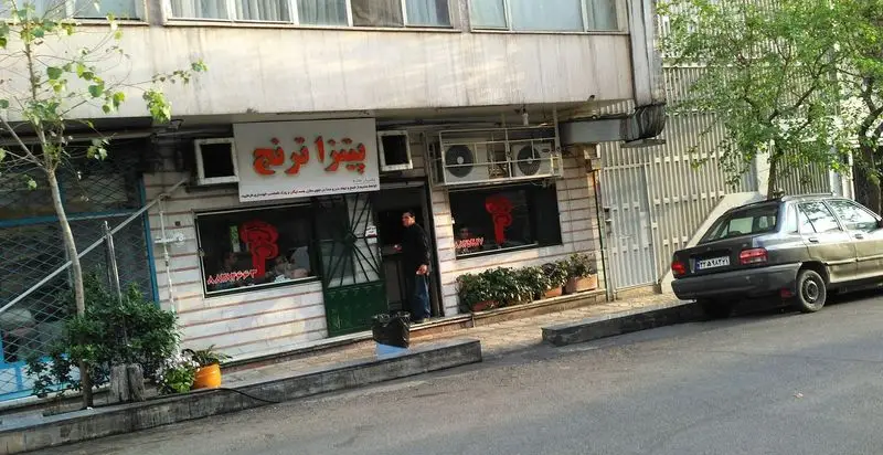 تصویری از نمای بیرونی فست‌ فود و پیتزا ترنج، تابلوی نارنجی رنگ رستوران با مردی در ورودی. شامل یک خودروی پارک شده و فضای سبز خیابان.