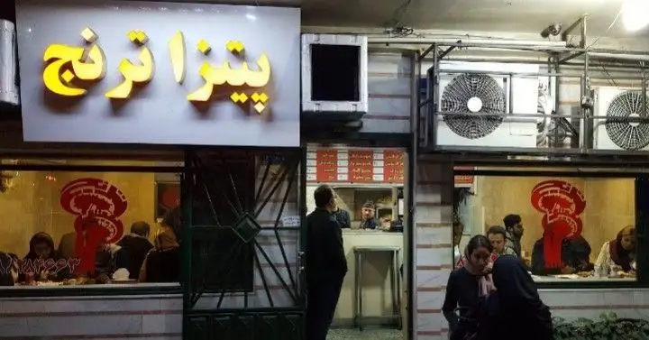 نمای شبانه فست‌فود پیتزا ترنج با تابلوی نئون روشن، مشتریان در حال صرف غذا و دکوراسیون داخلی متمایز. مکانی عالی برای تجربه غذای سریع.