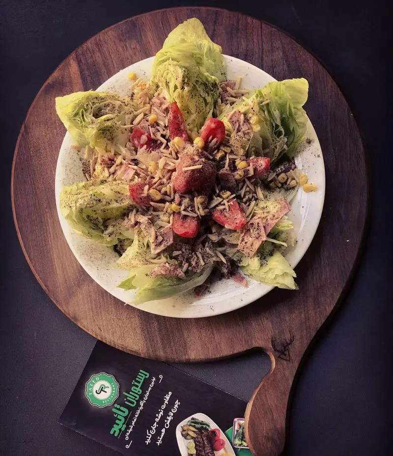 تصویری از یک سالاد تازه و اشتهاآور از رستوران تأیید، شامل کاهو، گوجه‌فرنگی و ذرت، که روی تخته چوبی سرو شده و بروشور منوی رستوران نیز پیداست.