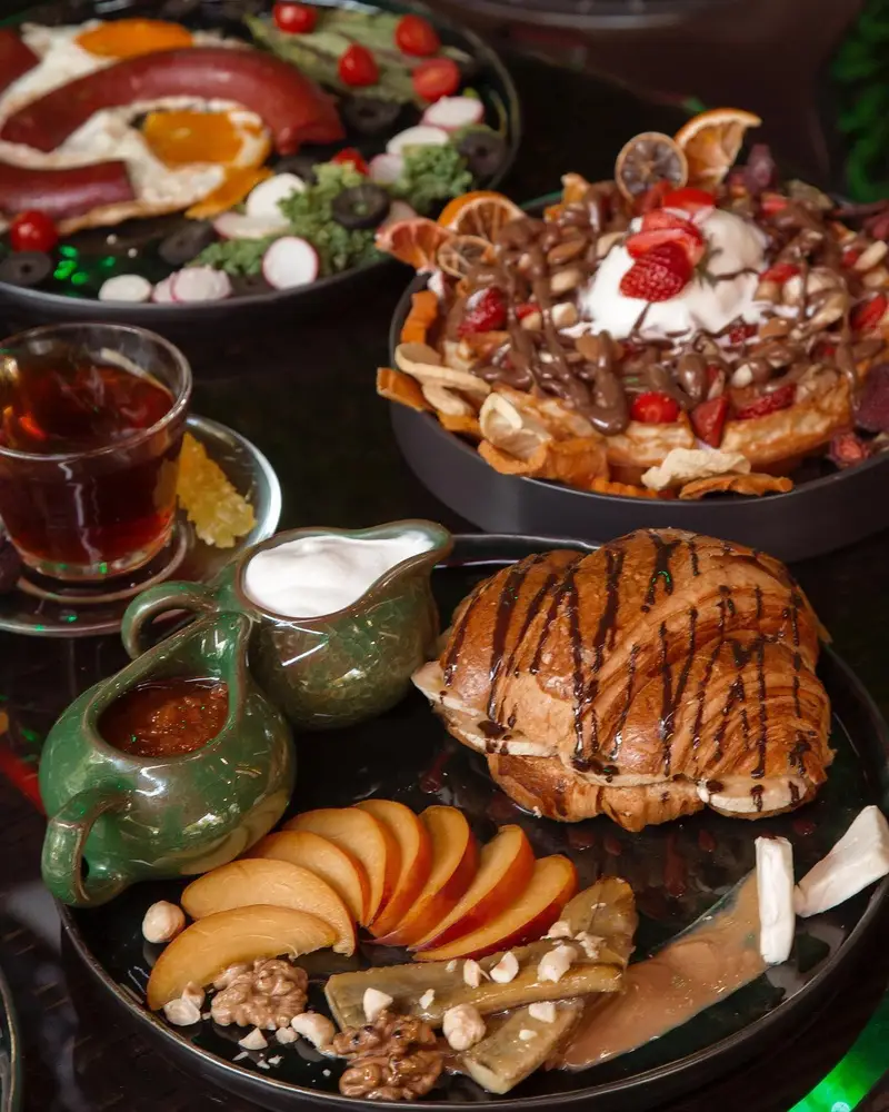 نمایی دلپذیر از میز صبحانه و برانچ مجلل کافه ماها شامل املت، سوسیس، وافل شکلاتی، کروسان، میوه و چای گرم.