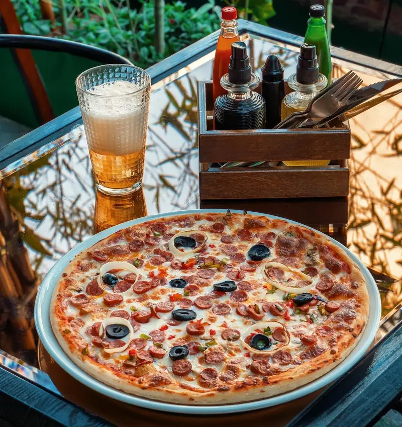 پیتزا سوسیس و زیتون خوشمزه کافه ماها روی میز براق در فضای دلنشین رستوران، همراه با لیوان آبجو خنک و سس‌های متنوع.
