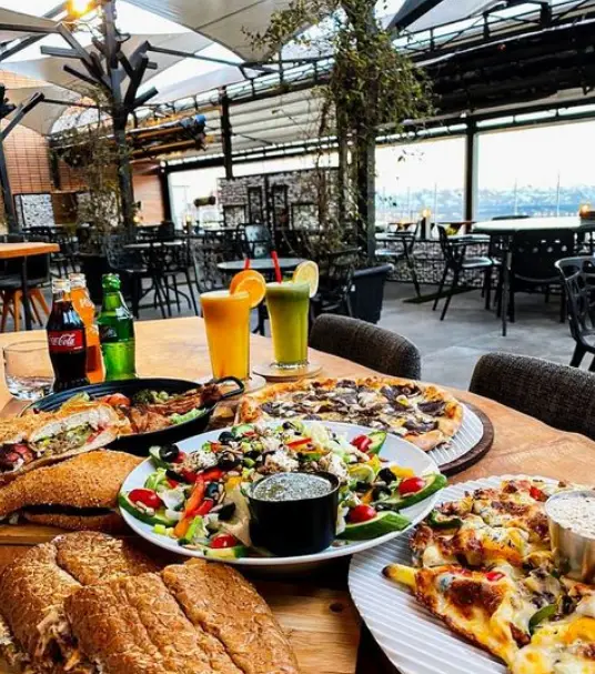 سفره رنگین غذا در رستوران فضای باز امیر شکلات دارآباد، شامل پیتزا، سالاد، ساندویچ و نوشیدنی‌های خنک در محیطی دلپذیر.