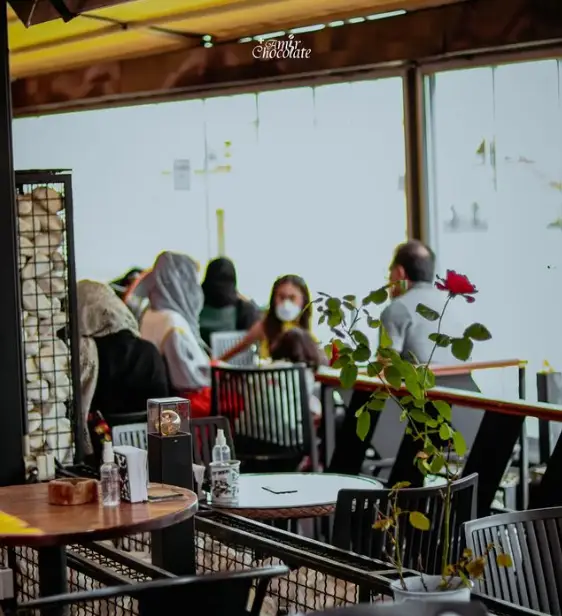 مشتریان در کافه امیر شکلات دارآباد، برخی با ماسک، در فضایی دلنشین نشسته و گپ می‌زنند، میزها با گلدان گل و اقلام بهداشتی.