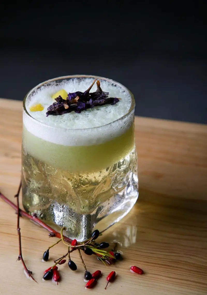 تجربه ای خاص در کافه رستوران رپتور: کوکتل خنک و دلپذیر با لایه های سبز و سفید، تزئین شده با گل‌های بنفش و بری‌های تازه، آماده سرو.