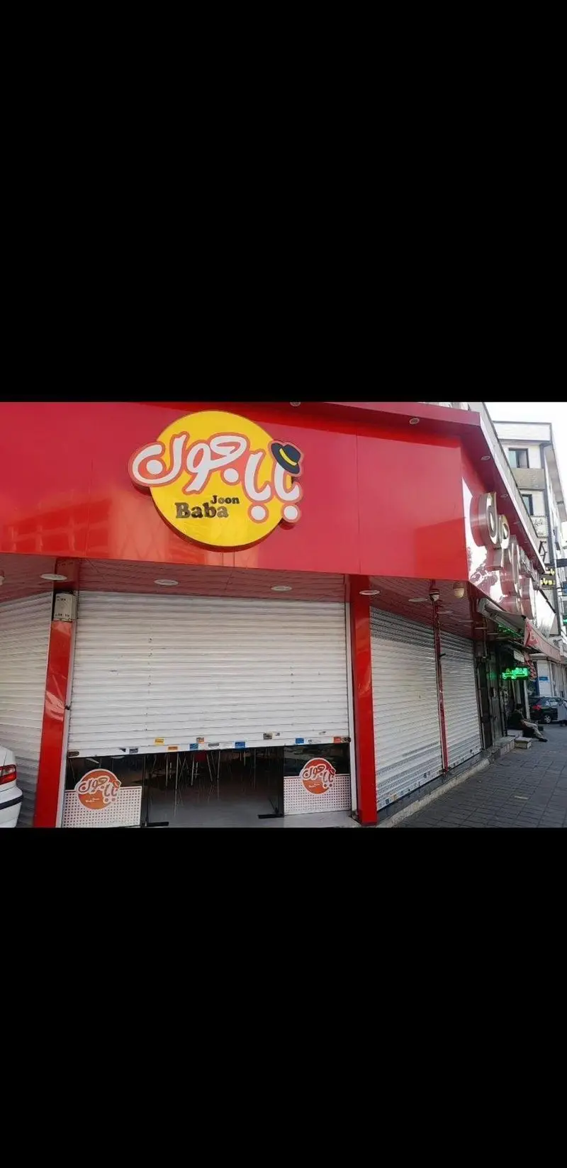 نمای بیرونی فست فود بابا جون نارمک با تابلوی بزرگ قرمز و زرد و آیکون کلاه، دارای کرکره‌های نیمه‌باز.