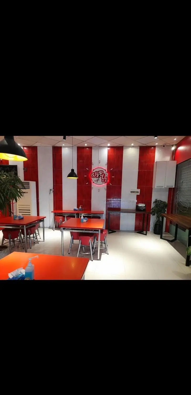 تصویر دکوراسیون داخلی شیک فست فود بابا جون نارمک، با طراحی دیوار قرمز و سفید، میزهای غذاخوری و ساعت دیواری نئون بابا جون.