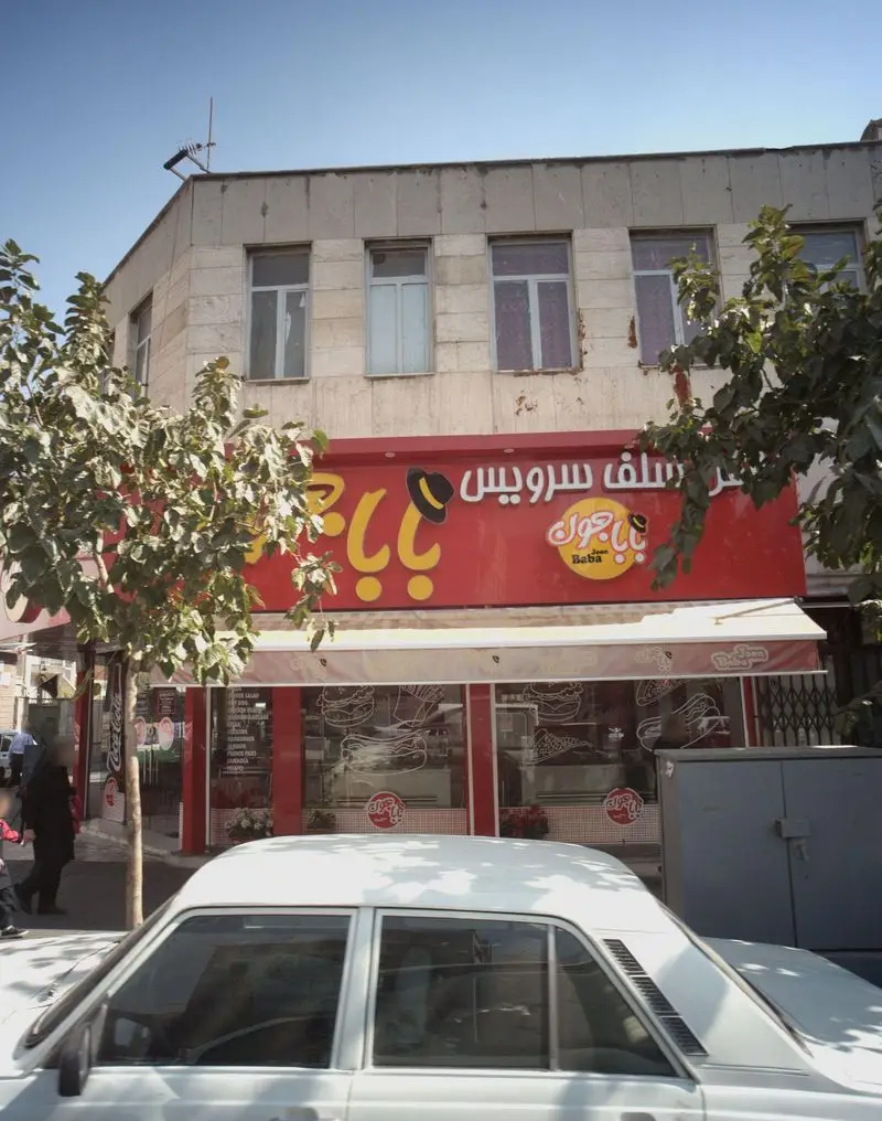 نمای بیرونی فست فود بابا جون نارمک با تابلوی قرمز رنگ، دکوراسیون سلف سرویس و تصاویر برگر، پیتزا و ساندویچ روی شیشه‌ها.