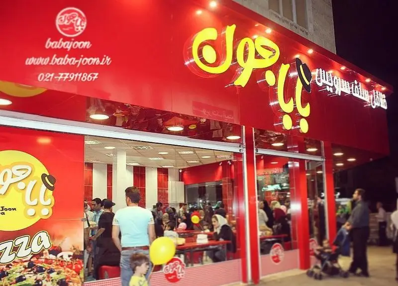 نمای خارجی فست فود بابا جون نارمک با تابلوی قرمز و زرد روشن، شامل لوگو و آدرس وبسایت، و تصویری از پیتزا. مشتریان در حال ورود و خروج.