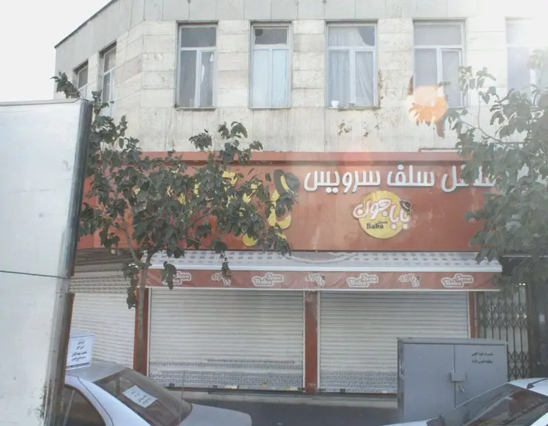 نمای بیرونی و تابلوی روشن فست فود سلف سرویس بابا جون در نارمک تهران، با لوگوی جذاب و کرکره‌های بسته مغازه.