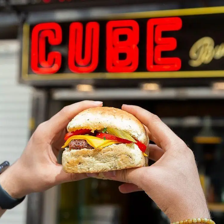 برگر خوشمزه کیوب برگر با پنیر و سبزیجات تازه در دست، با تابلوی نئون قرمز CUBE در پس‌زمینه. تجربه غذایی لذیذ