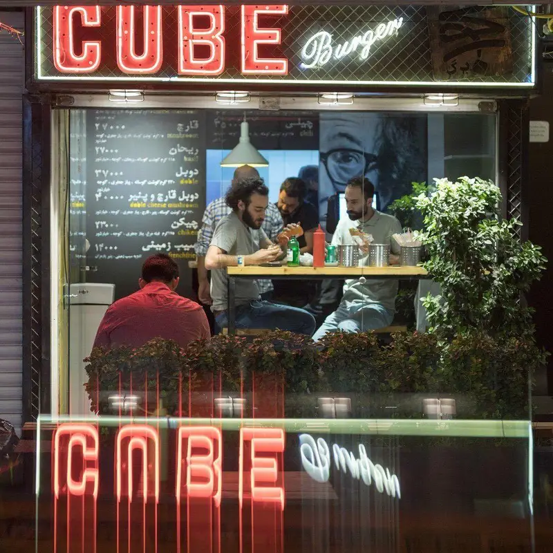 تصویری از نمای شب یک رستوران فست فود شیک "کیوب برگر" با مشتریان در حال لذت بردن از همبرگر در فضای داخلی روشن و تابلوهای نئون قرمز.