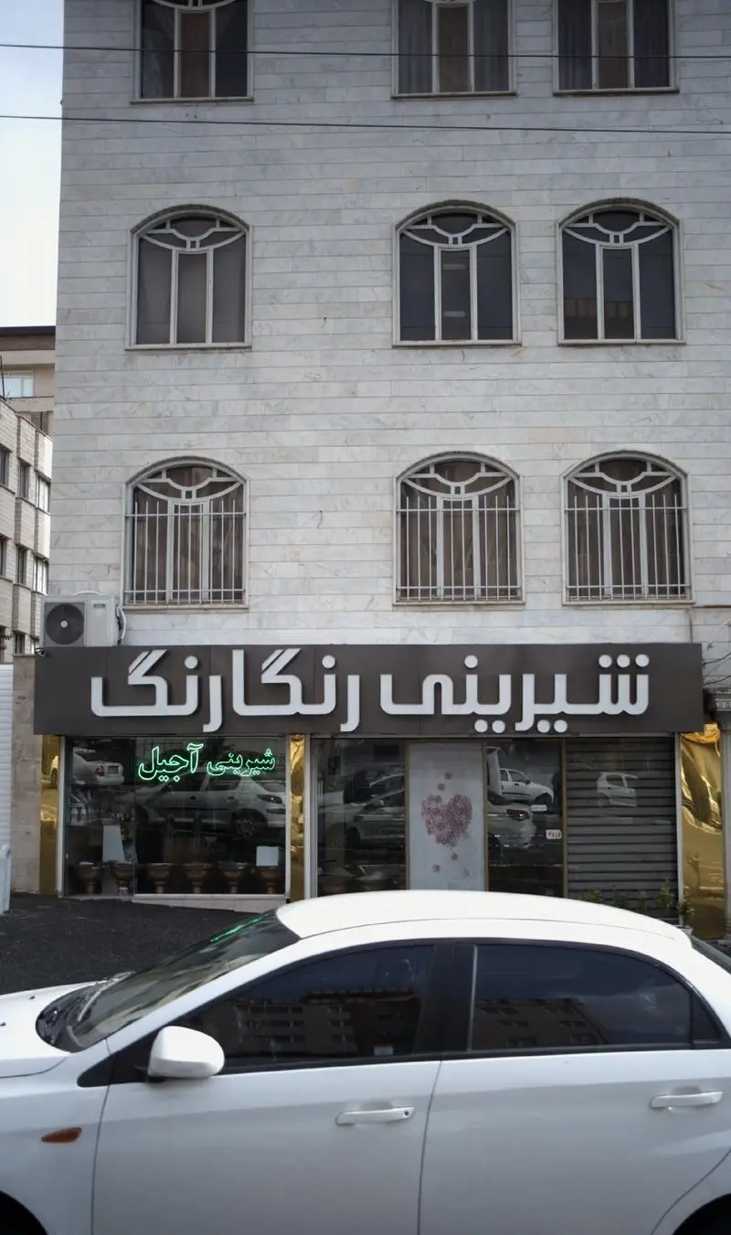 تصویر نمای بیرونی فروشگاه شیرینی رنگارنگ با تابلوی بزرگ و شیشه‌های رفلکتیو، مکانی برای خرید شیرینی و آجیل تازه.