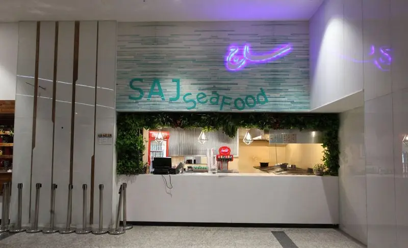 نمای داخلی فود کورت ایران مال، رستوران ساج سی‌فود با پیشخوان سفارش غذای دریایی و دکوراسیون مدرن سبز و نور نئون.