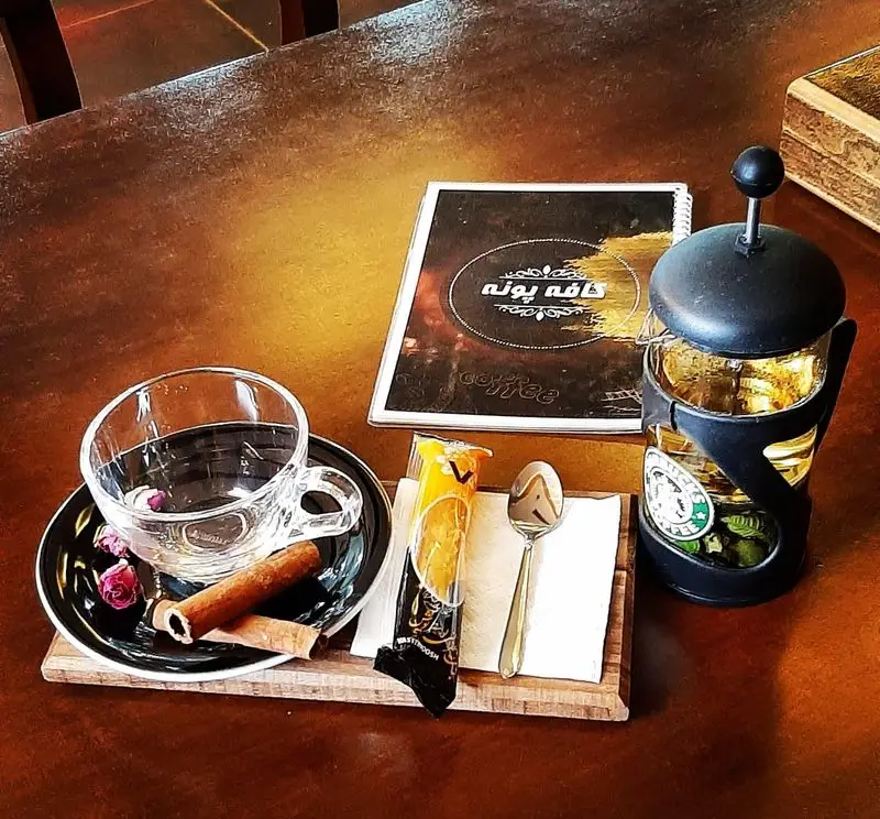 میز سرو یک نوشیدنی گرم و دلپذیر در کافه پونه: فرنچ پرس، فنجان، دارچین، گل رز، شیرینی و منوی جذاب کافه.