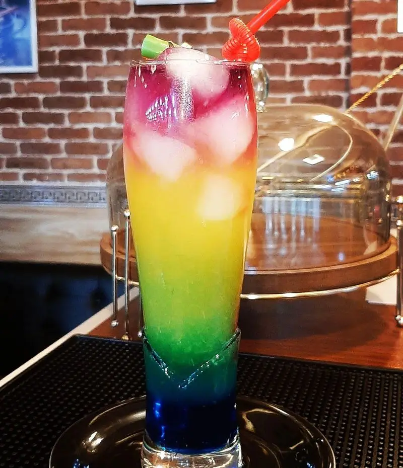 کوکتل رنگارنگ و لایه ای کافه پونه، نوشیدنی تابستانی خنک و جذاب با یخ و نی قرمز، در فضایی دلنشین و خاص.