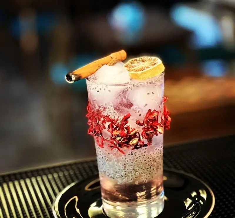 نوشیدنی رنگارنگ و خنک کافه پونه، شامل دمنوش هیبیسکوس، بذر ریحان، یخ، لیمو و چوب دارچین دودی شده، انتخابی دلپذیر.