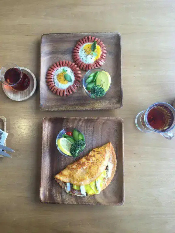 نمایی از صبحانه کامل و لذیذ کافه بارن: املت تازه، سوسیس تخم مرغ با تزئین گل و سبزیجات متنوع به همراه چای داغ روی میز چوبی.