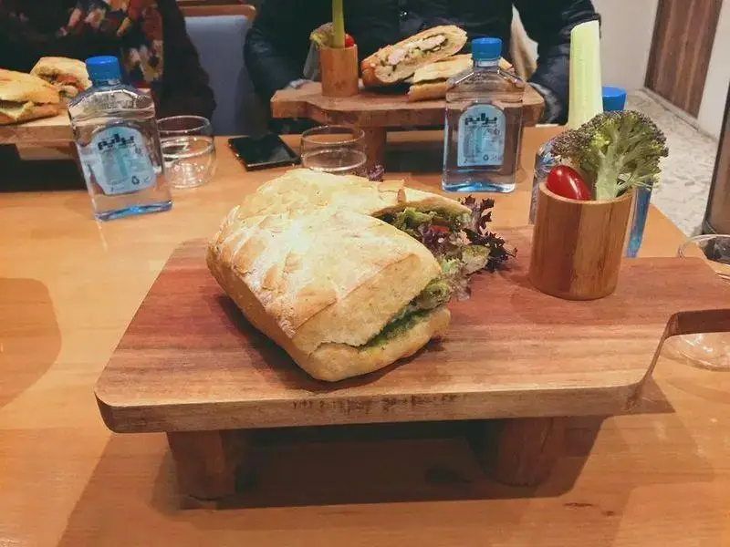 ساندویچ بزرگ و خوشمزه کافه بارن روی تخته چوبی، همراه با سالاد، سبزیجات تازه و آب معدنی پراو روی میز چوبی رستوران.