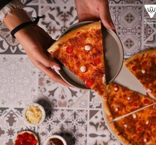 دست در حال سرو اسلایس پیتزا پیتزا سیب ۳۶۰ با پنیر، گوجه گیلاسی و مینی موزارلا روی میز کاشی‌کاری شده با سس‌های متنوع.