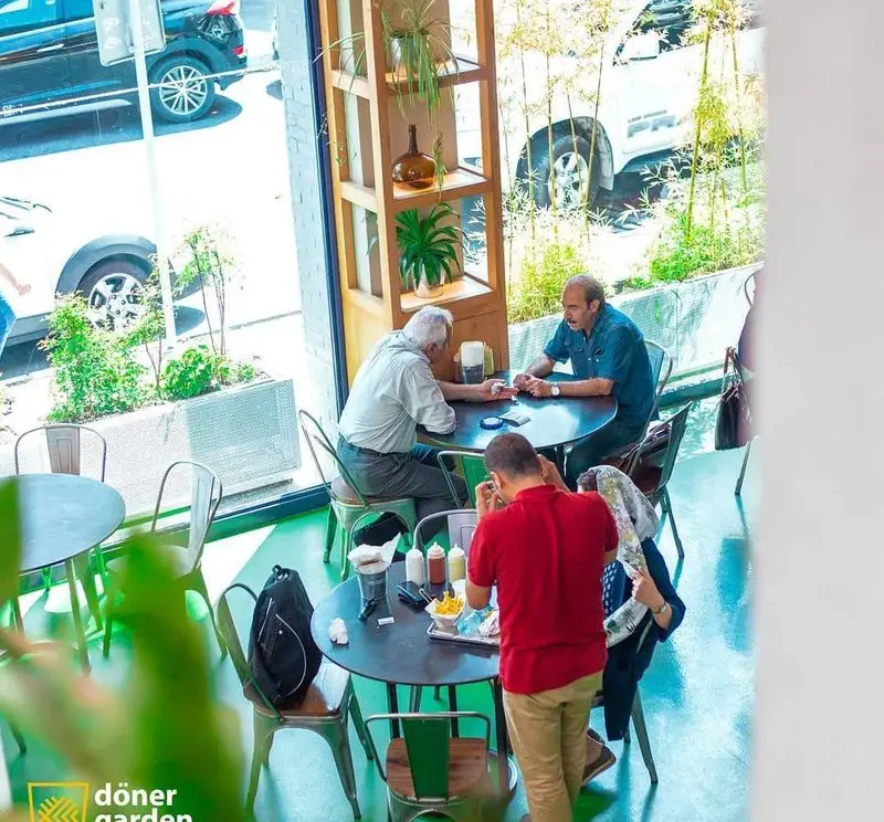 نمای دلنشین از فضای داخلی رستوران دونر گاردن با مشتریانی که در حال صرف غذا و گفتگو هستند. پنجره‌ای بزرگ رو به خیابان و دکوراسیون گیاهی زیبا.