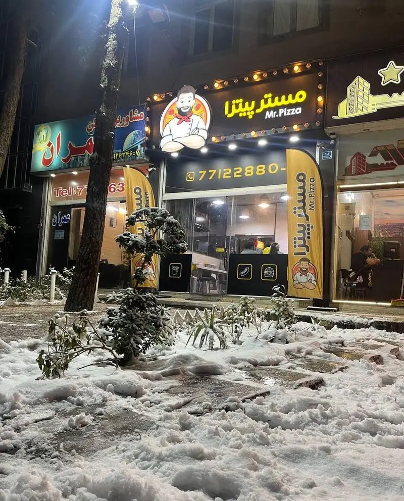 تصویری از نمای بیرونی کسب و کار مستر پیتزا در شب برفی زمستانی، با تابلوی روشن، لوگوی سرآشپز و شماره تلفن 77122880.