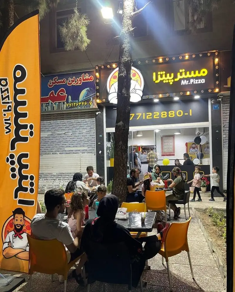 مستر پیتزا: رستورانی شلوغ در شب با خانواده‌ها در فضای باز، تابلوی نئونی درخشان و بنر تبلیغاتی سرآشپز پیتزا.