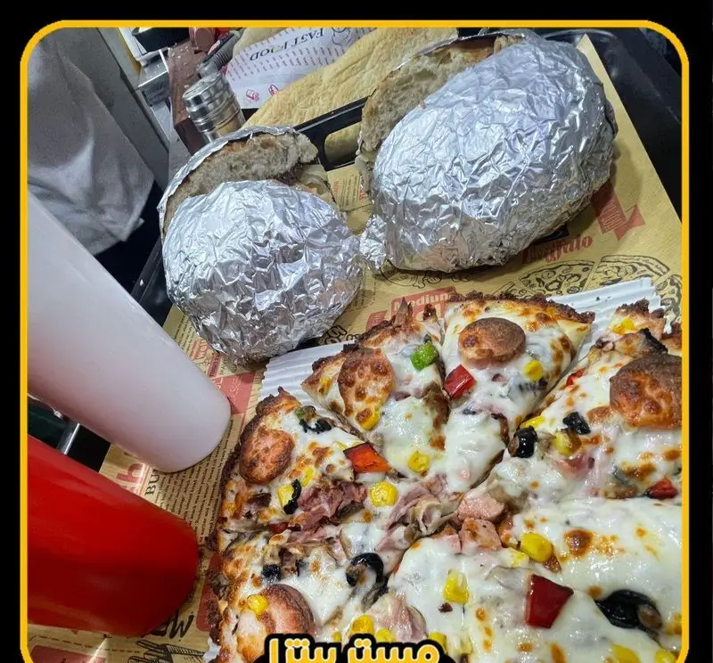 تصویری از پیتزای عالی مستر پیتزا با پنیر فراوان، سوسیس، فلفل دلمه‌ای، ذرت و زیتون. دو ساندویچ فویلی و سس‌های قرمز و سفید نیز کنار غذا قرار دارند.