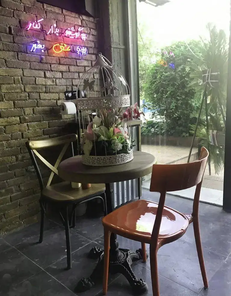 فضای دنج و دلنشین کافه چارچیز با میز و صندلی راحت، دیوار آجری، تابلوی نئون، قفس دکوری و منظره طبیعت از پنجره.