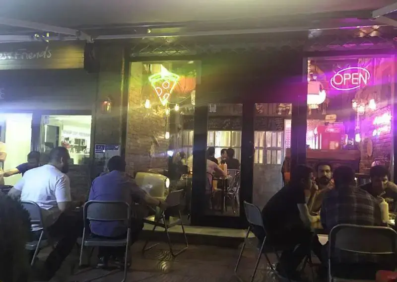 نمایی شبانه از رستوران و کافه چارچیز، مملو از مشتریان در فضای باز، با تابلوهای نئون پیتزا و چراغ 'OPEN' که جلوه‌ای خاص به آن داده است.