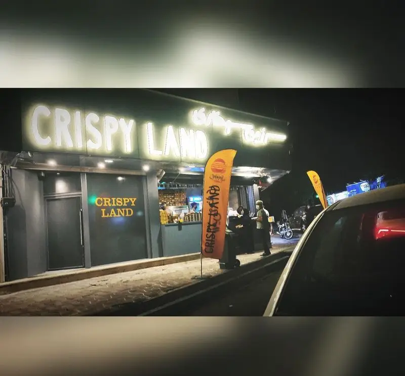 تصویر شبانه از نمای بیرونی فست فود سرزمین سوخاری هروی با تابلوهای درخشان و پرچم‌های نارنجی Crispy Land.