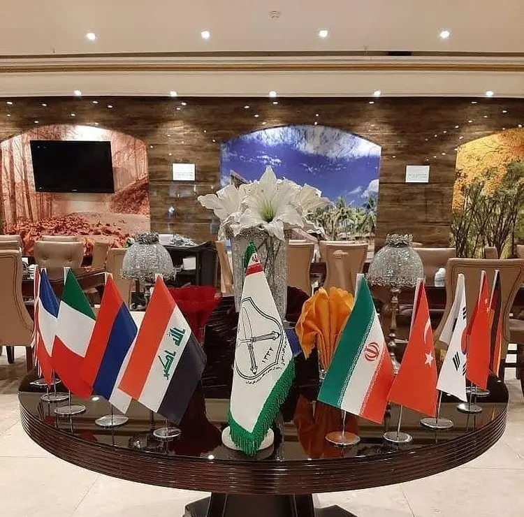 میز گرد زیبا در رستوران آرامیس، مزین به پرچم‌های ملی و گلدان گل سفید، نشان‌دهنده محیطی لوکس و بین‌المللی برای مهمانان.