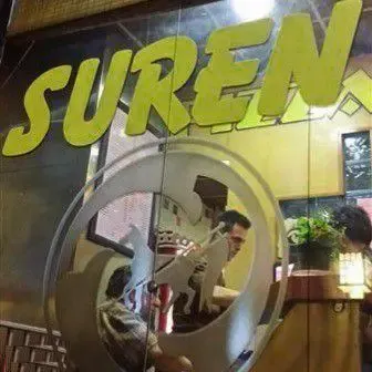 نمایی از درب ورودی شیشه‌ای پیتزا سورن با لوگوی بزرگ و برجسته "SUREN" به رنگ زرد، منعکس‌کننده فضای داخلی گرم و دوستانه رستوران فست فود.
