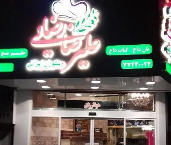 نمای بیرونی شبانه کبابی علیرضا رضایی، تابلوی نئون درخشان با لوگوی سرآشپز و شاخه‌های زیتون. ارائه نان داغ و کباب داغ. شماره تماس ۷۷۲۳۰۰۲۳.