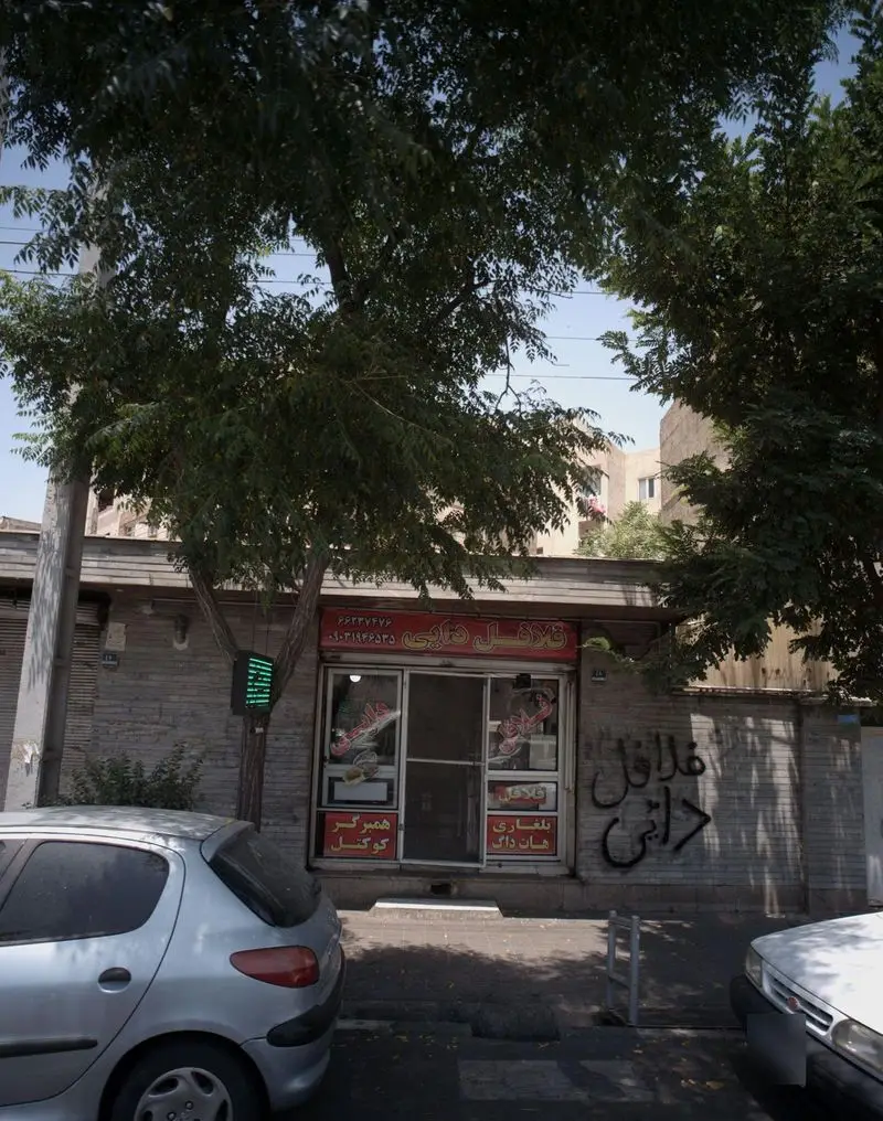 نمایی از مغازه فلافل دایی در خیابان، با تابلو قرمز، شیشه‌های ویترین، دو اتومبیل در جلو و درختان سبز. خدمات فلافل، همبرگر، هات داگ.