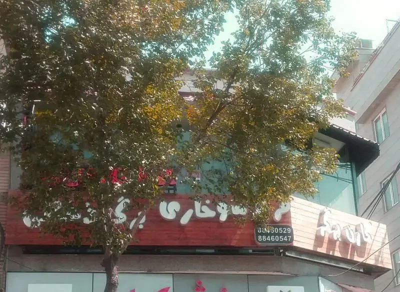 نمایی از فست فود کارن برگر با نمای چوبی، تابلوی بزرگ با نوشته فارسی و درخت سبز در مقابل آن، همراه با شماره تلفن.